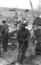 Amongst these rig workers in the field, ca.1982, some are wearing hardhats, others ordinary baseball caps, and one even smokes a cigarette.