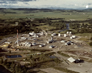 In this aerial view of the Turner Valley gas plant, ca. 1985, the sulfur plant and attached power plant are just to the right of the tall red and white stack. Note the stockpile of yellow sulfur at left. <br />Source: Alberta Culture and Tourism