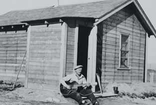 An oil worker plays his guitar outside of a bunkhouse, 1938